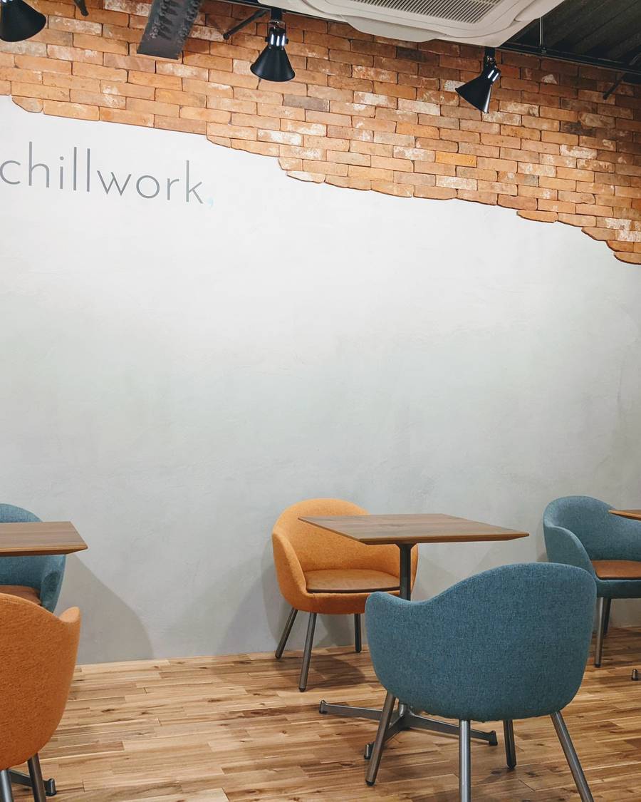レンガと漆喰の壁にchillworkのロゴ。木の机と青緑・オレンジの椅子が並ぶフリースペース