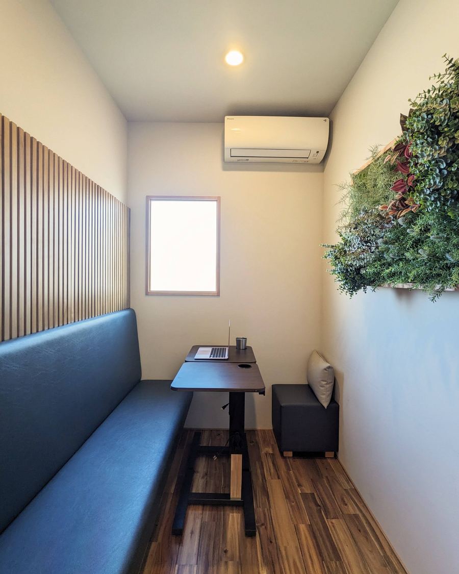 プレミアムの個室P-02。奥行きのある部屋に長いソファと机、壁に植物のパネル、窓とエアコン